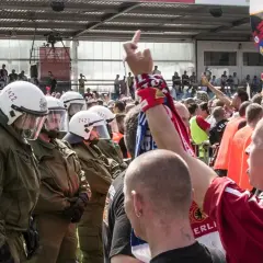 Policja w pełnym rynsztunku stoi naprzeciwko grupy kibiców, wśród których widać ultrasi z flagami i szalikami.