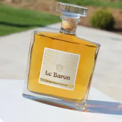 Bouteille de "Le Baron Grande Réserve Gérard", un spiritueux ambré, posée sur un socle blanc, avec un arrière-plan flou évoquant un jardin et un chemin, rappelant l'élégance d'un chateau vaudois.