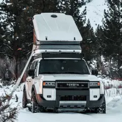 Biały samochód Toyota z rozłożonym namiotem dachowym w zimowym lesie. Pokazano, jak zrobić namiot dachowy, rozkładając drabinę.