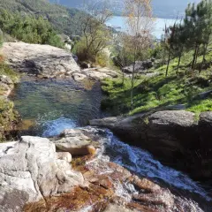 Cascada y pozas naturales en Pozas da Moura, con agua cristalina cayendo sobre rocas y vegetación exuberante. Al fondo, un estuario y colinas verdes.