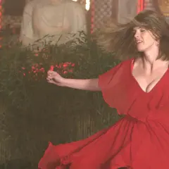 En kvinna i röd klänning dansar. Bakom henne syns en staty och bambu. Scen från "three women serie".