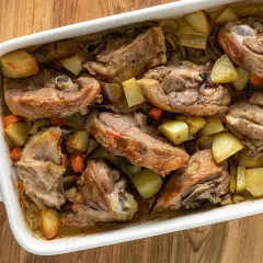Capretto al forno ricetta della nonna: succulenti pezzi di carne dorata con patate e carote in una teglia bianca.