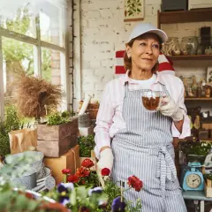 Frau mit Kappe und Schürze trinkt Tee im Blumenladen. Sie genießt ihre Zeit, vielleicht im Rahmen von altersteilzeit modellen, umgeben von Pflanzen.