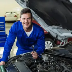 Uśmiechnięty mechanik w niebieskim kombinezonie z kluczem w dłoni, pracujący przy silniku. Dowiedz się, jak zostać elektrykiem samochodowym.