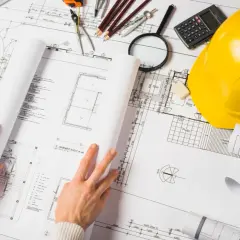 Manos revisando planos de construcción, con casco amarillo, calculadora y lupa. La ite del proyecto se detalla en los planos.