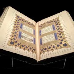 En öppen, gammal Koran på ett ställ, med detaljerad kalligrafi och gyllene ornament. En påminnelse om vem som skrev Koranen.