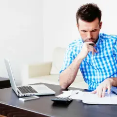 Mężczyzna w kratę analizuje dokumenty, obok laptop, telefon i kalkulator. Może to być jego ufg historia oc.