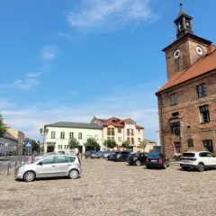 Rynek w Połajewie z zabytkowym ratuszem i kolorowymi kamienicami. Na brukowanym placu zaparkowane samochody.