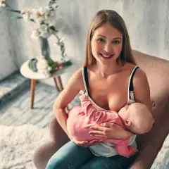 Uśmiechnięta mama karmi piersią dziecko w pozycji klasycznej do karmienia, siedząc w fotelu.