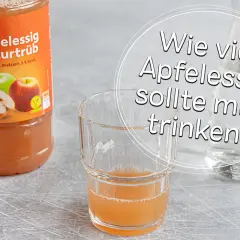 Flasche naturtrüber Apfelessig neben einem Glas mit Apfelessig-Wasser-Mischung. Die Frage "Wie viel Apfelessig sollte man trinken?" wird thematisiert.