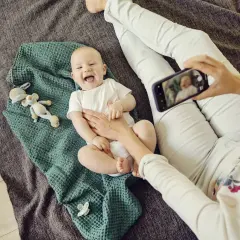 Uśmiechnięte niemowlę do kiedy będzie tak radosne? Mama robi mu zdjęcie telefonem, leży na kocyku z zabawkami.