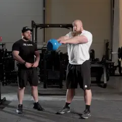 Två män i gym utför kettlebell swing. Den ena mannen, med skägg, håller i en blå kettlebell och är i rörelse. Den andra mannen står bredvid och observerar.