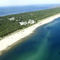 Jurata plaża z lotu ptaka: szeroka, piaszczysta plaża, sosnowy las i budynki hotelowe nad turkusowym morzem.