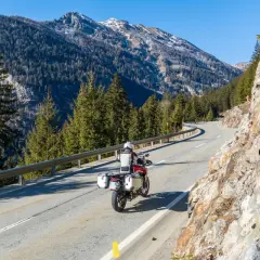 Un motociclista affronta una curva su una strada di montagna, godendosi un perfetto week end in moto tra boschi e cime innevate.