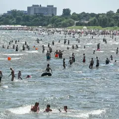 Tłum ludzi kąpiących się w Bałtyku, na tle plaży z parasolami i budynków.