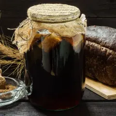 Słoik z ciemnym napojem, obok bochenek chleba żytniego i miseczka z mąką – składniki do domowego kwasu chlebowego.