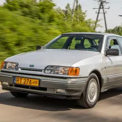 Srebrny Ford Scorpio jedzie po drodze, otoczony zielenią.