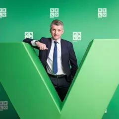 Mężczyzna w garniturze opiera się o zieloną literę "V" z logo Velo Bank.