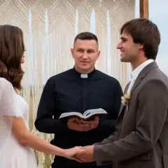 Ksiądz na ślubie, trzymając Biblię, błogosławi parę młodą. Panna młoda w białej sukni i welonie, pan młody w garniturze.