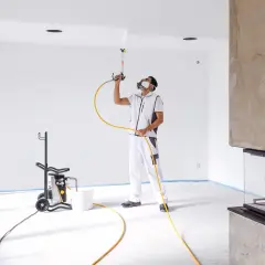 Pittore con maschera protettiva usa una pistola per vernice a spruzzo per dipingere il soffitto di una stanza vuota.