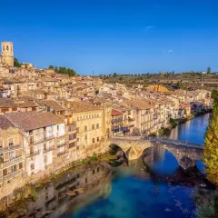 Pueblo con casas de tejas de barro y un puente de piedra sobre un río azul, evocando la toscana española.