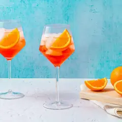 Två glas med Aperol Spritz, is och apelsinskivor, med en hel apelsin och skivor på en skärbräda i bakgrunden.