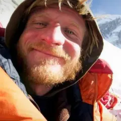 Uśmiechnięty alpinista z zarostem, w kolorowej kurtce, na tle ośnieżonych gór. W tle inna osoba w czerwonej kurtce. Może to być objaw choroby wysokościowej.