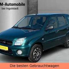 Zielony Subaru Justy z żółtą tabliczką "TOP ZUSTAND" na tle hali dealerskiej.