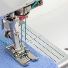 Close-up of a sewing machine needle plate and foot, demonstrating how to use a double needle on blue fabric with turquoise thread.