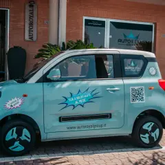 Macchina piccola 4 posti azzurra, con adesivi promozionali "TUA A 99,00 euro AL MESE" e QR code.