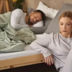 Mujer triste sentada en el suelo, mientras su pareja duerme en la cama. Reflexiona sobre qué significa soñar que tu pareja te es infiel.