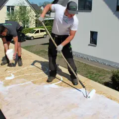 Zwei Handwerker arbeiten auf einem Dach mit OSB-Platten. Einer gießt weiße Flüssigkeit aus einem Eimer, der andere verteilt sie mit einer Rolle.