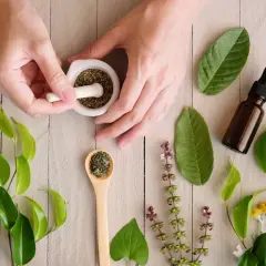 Hände verarbeiten Kräuter im Mörser, um die adaptogene Wirkung zu nutzen. Daneben liegen Öl-Kapseln, getrocknete Kräuter und eine Pipettenflasche.
