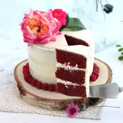 En vacker red velvet cake med fluffig frosting, dekorerad med hallon och rosa blommor. En bit skärs upp.
