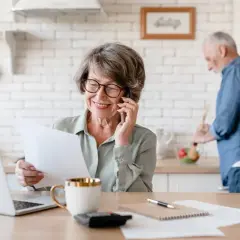 Uśmiechnięta kobieta rozmawia przez telefon, trzymając dokumenty. Obok laptop, filiżanka i kalkulator. Mąż gotuje w tle. 47 lat pracy jaką emerytura.