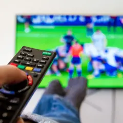 Dłoń trzymająca pilot do telewizora, na ekranie mecz piłkarski.