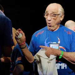 Gene Lebell, med förvånat uttryck, håller upp en tidtagningsklocka och pekar.