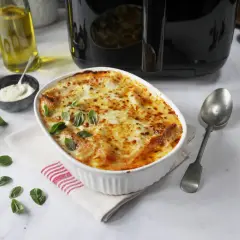En gyllene lasagne i airfryer, toppad med smält ost och färsk basilika. Serveras med en sked och tillbehör som olivolja och parmesan.