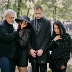 Smutna rodzina na cmentarzu. Elegancki ubiór damski na pogrzebie zimą, z kapeluszem i okularami przeciwsłonecznymi, obok żałobników w czerni.