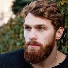 Un homme au visage ovale, avec une barbe rousse et des yeux bleus perçants, regarde au loin, entouré de verdure.