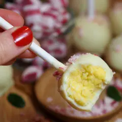 En hand med rött nagellack håller en cake pop med vit choklad och krossade polkagrisar. Perfekt för ett cake pops recept!