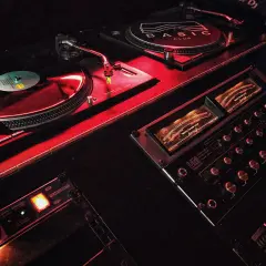 Giradischi e mixer in un locale notturno, illuminati da luci rosse. Il logo "BASIC CLUB" è visibile su uno dei giradischi.