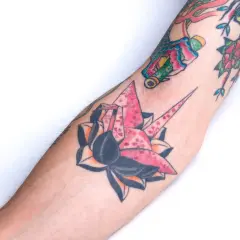 Kolorowe tatuaże na ramieniu: różowy żuraw origami z kwiatami wiśni, róża i inne wzory.