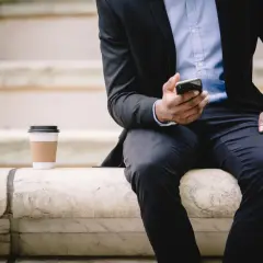 Mężczyzna w garniturze siedzi na kamiennych schodach, trzymając telefon. Kawa obok. Czeka na ważne powiadomienie, może to być przekierowanie na nowe zadanie.