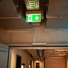 Zielone oświetlenie awaryjne z ikoną biegnącej postaci i strzałką w dół, zgodne z przepisami, wskazuje drogę ewakuacyjną.