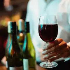 Main d'un homme tenant un verre de vin rouge, prêt pour une dégustation de vin Beaujolais. Des bouteilles floues en arrière-plan.