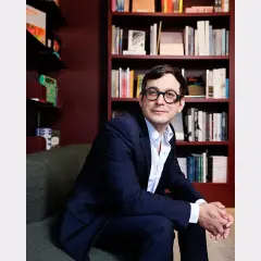 A man in a suit and glasses sits in front of bookshelves, perhaps at the David Lewis Gallery, looking thoughtfully to the side.
