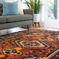 Ein detailreicher perserteppich mit roten und blauen Mustern liegt vor einem grauen Sofa. Tipps zum perserteppich reinigen.
