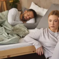 Eine Frau sitzt nachdenklich am Bettrand, während ihr Partner schläft. Sie kämpft mit ihrer Sexsucht bei Frauen.