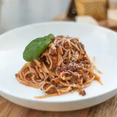 Porcja spaghetti bolognese z parmezanem i listkiem bazylii na białym talerzu.